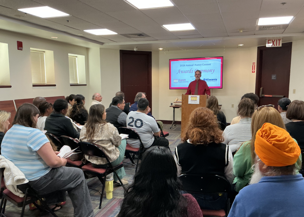 The library director stands behind a podium addressing a crowd in the library meeting room.